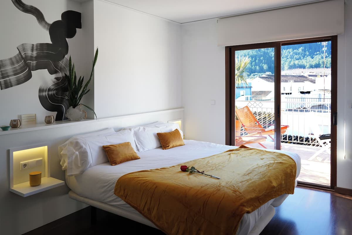 Una habitación de hotel moderna con una cama grande, una mesita de noche con lámpara, vistas de la ciudad a través de un ventanal amplio y un elemento decorativo en la pared.