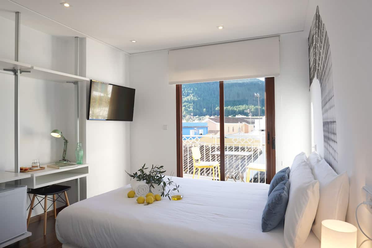 Un hotel moderno con una cama grande, un escritorio con silla, televisor de pantalla plana y un balcón con vistas a la ciudad y a las montañas.