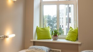 A bedroom with a window, two green pillows, a potted plant, and a bed with a patterned blanket. - Liberty Airy (Riga)
