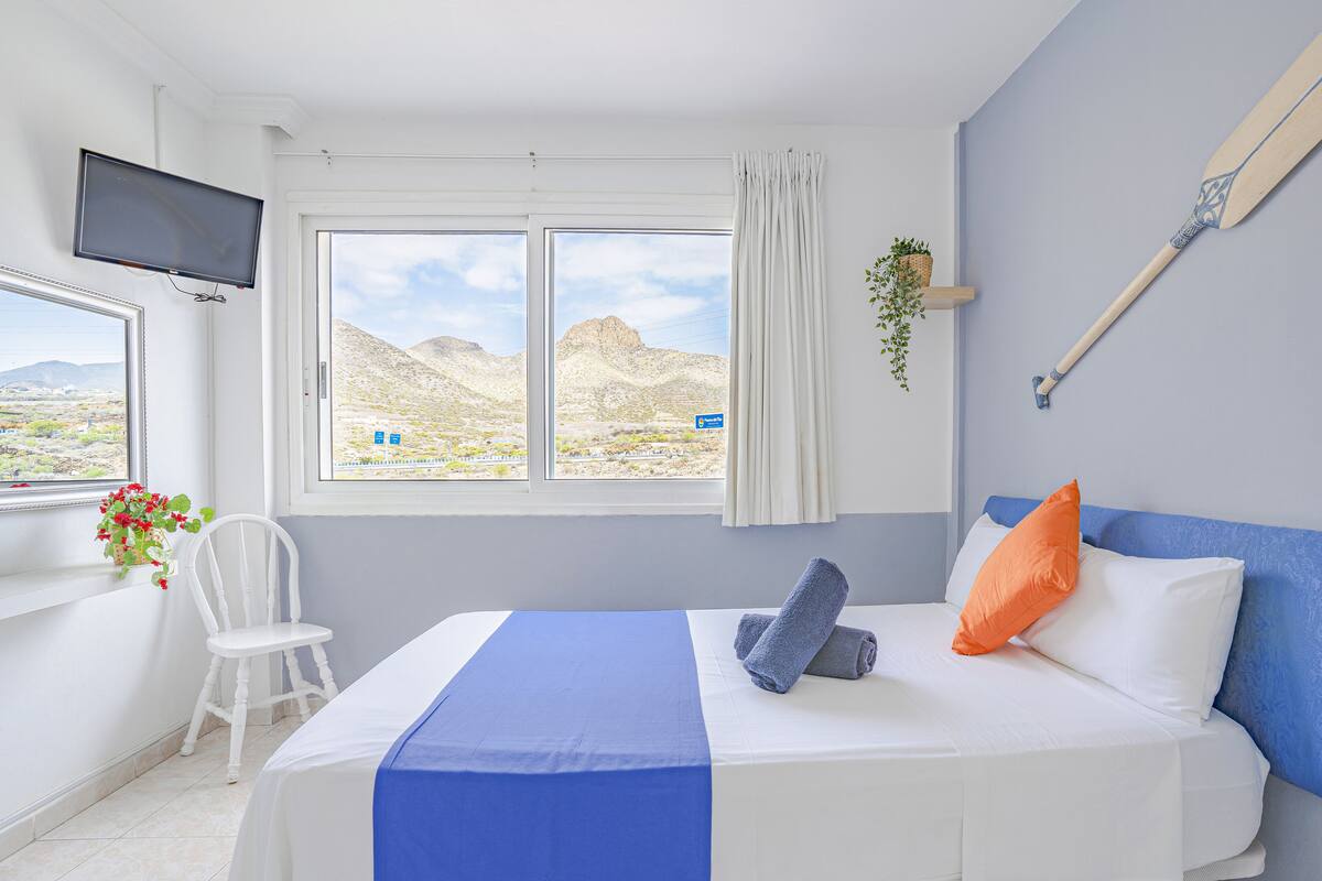 Quarto de hotel com cama, televisão, janela com vista para o deserto, cadeira e planta.