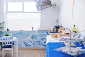 A room with a blue and white wall mural, a white table with a blue tablecloth, a white chair, and a buffet table with food items. - Hostel Neon Tenerife (Arona)
