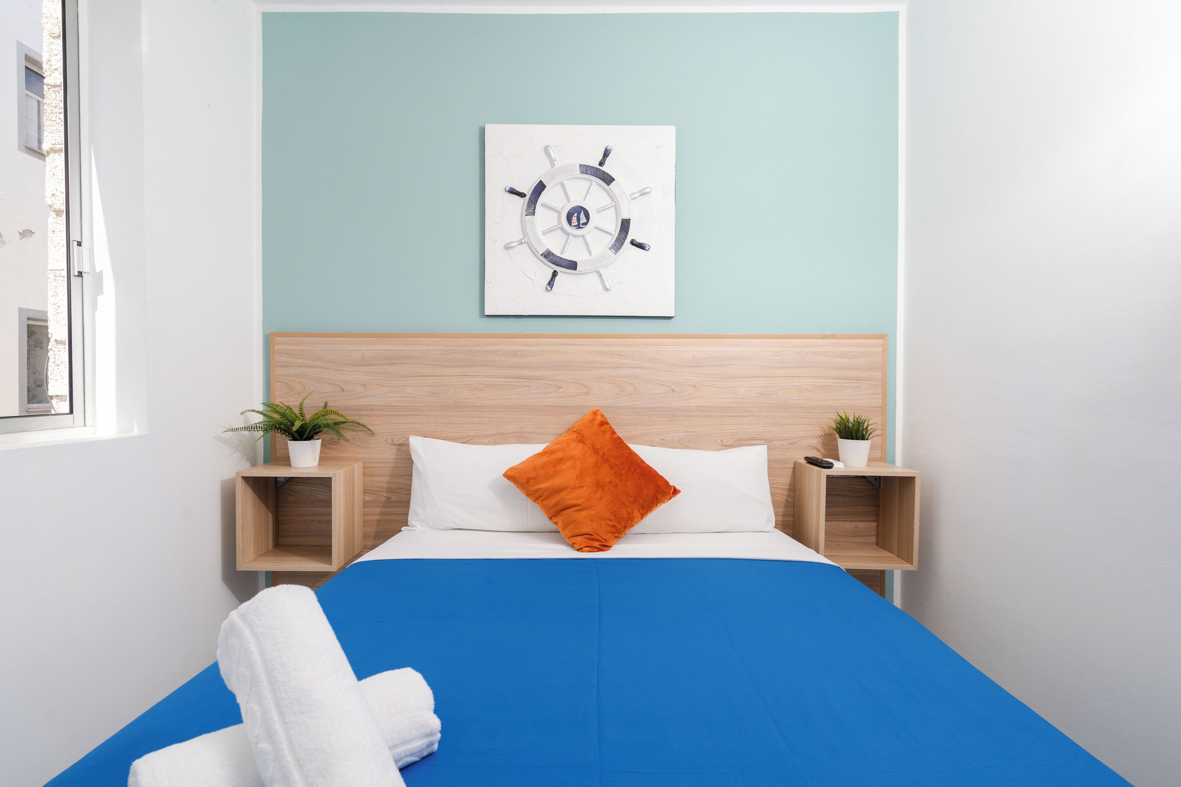 A modern hotel room with a wooden headboard, a blue bedspread, white pillows, and a nautical-themed wall art.