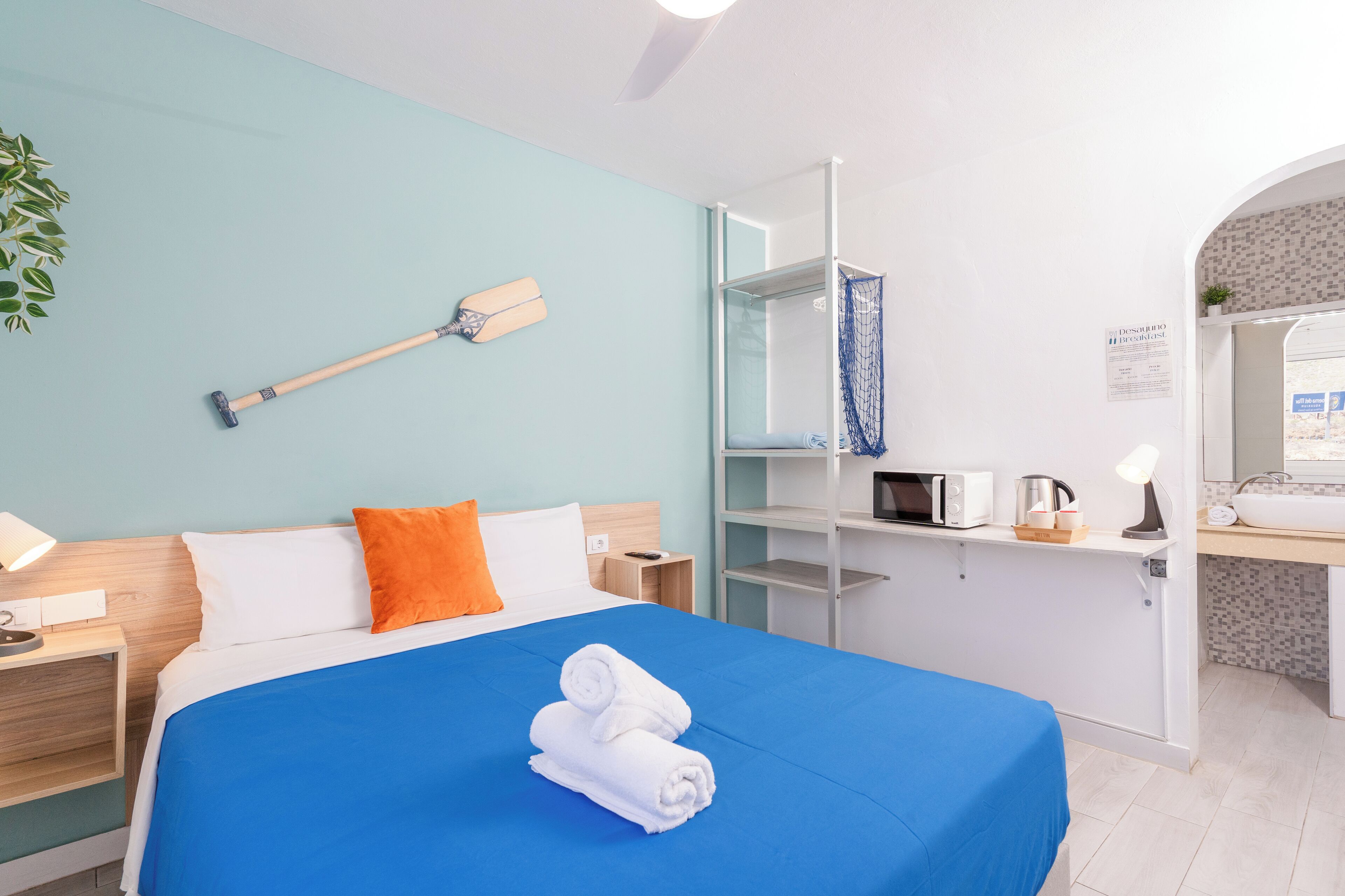 A modern hotel room with a bed, a desk, a microwave, a kettle, and a small plant.