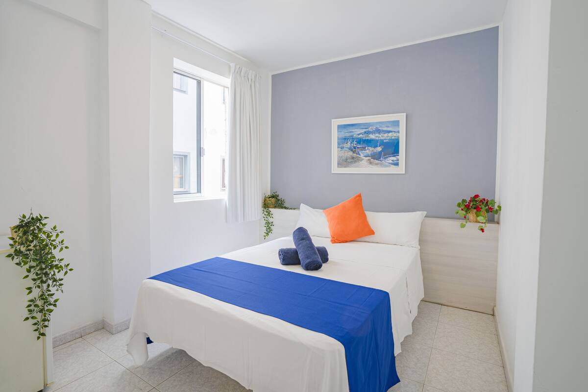 Quarto de hotel com cama, janela com cortinas, um quadro na parede e uma planta no criado-mudo.