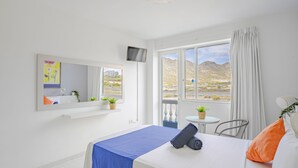 A hotel room with a bed, a TV, a window with a view of the mountains, and a small table with a plant.