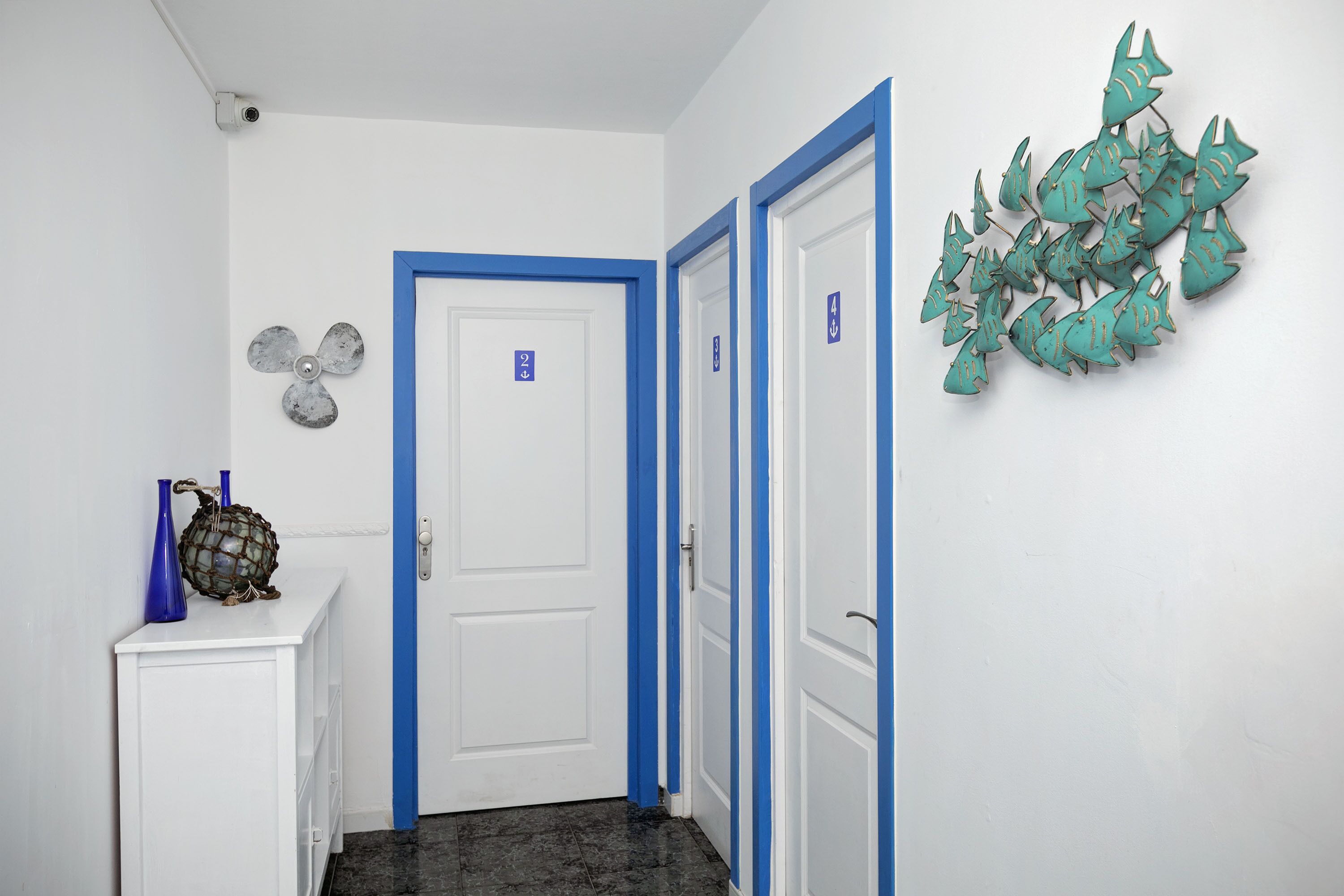 Un couloir aux murs blancs, avec des moulures bleues et une porte blanche dotée d’un cadre bleu. Un élément décoratif orne le mur de droite, et un petit meuble avec des vases bleus se trouve à gauche.