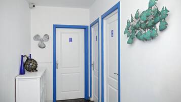 Un couloir aux murs blancs, avec des moulures bleues et une porte blanche dotée d’un cadre bleu. Un élément décoratif orne le mur de droite, et un petit meuble avec des vases bleus se trouve à gauche.