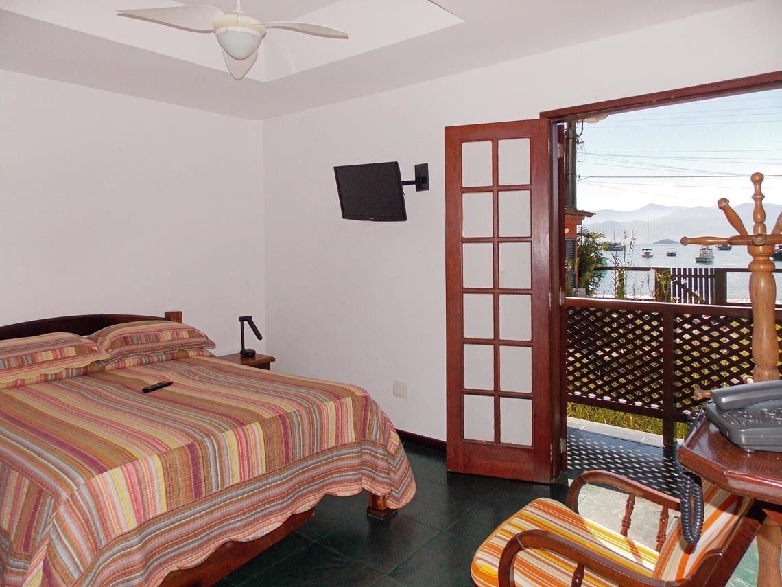Un dormitorio con cama, una silla, un escritorio con teléfono, televisión montada en la pared y un balcón con vistas a barcos y montañas.