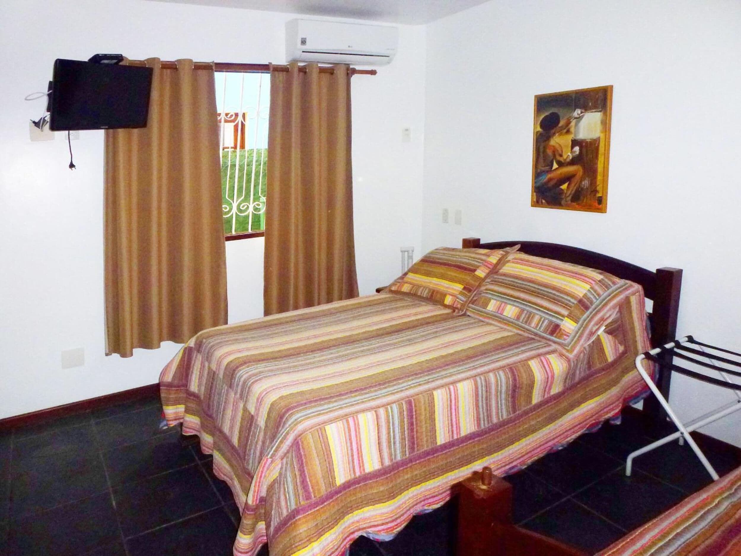 A bedroom with a bed, curtains, a painting, and an air conditioning unit.