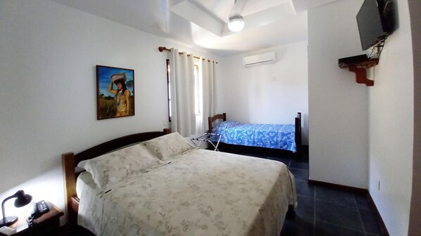 A bedroom with a large bed, a smaller bed, a desk with a lamp, a painting on the wall, and an air conditioning unit.
