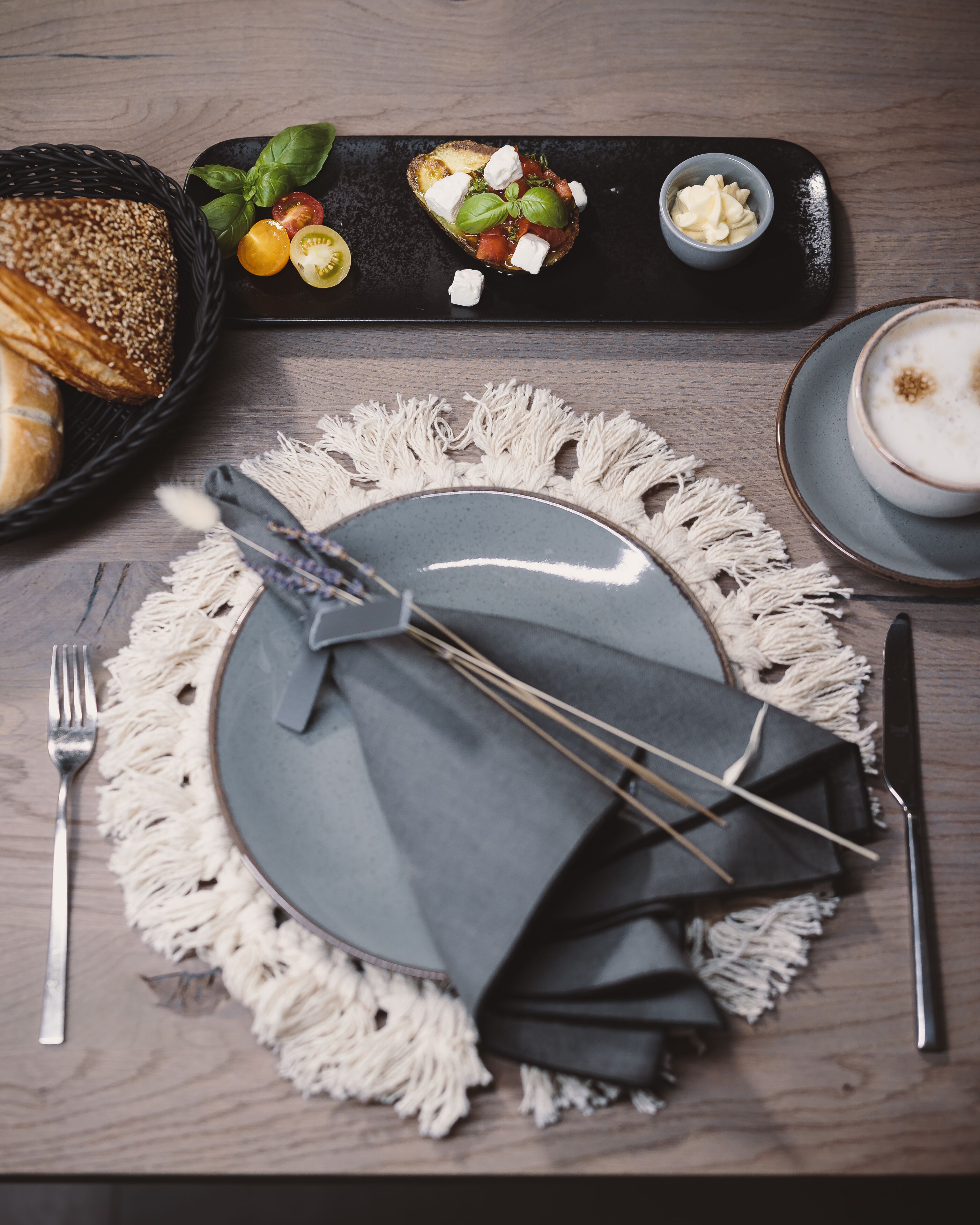Een eettafel gedekt met een bord, bestek, een servet, een schaal kaas en een mand brood.