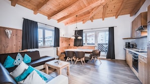 A cozy living room with a wooden ceiling, a sofa with patterned cushions, a dining area with a table and chairs, and a kitchen with wooden cabinets and a stove. - Margarethenstein Boutique Hotel & Apartment (Kaprun)