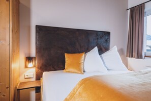Habitación de hotel con una cama grande, cabecera de madera, mesita de noche con lámpara y ventana con cortinas.