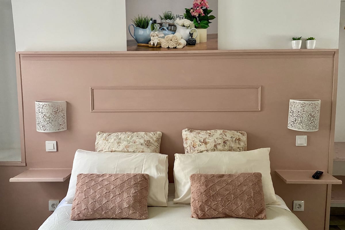 A bedroom with a bed, two pillows, a nightstand, a wall-mounted lamp, and a decorative shelf with plants and a vase.