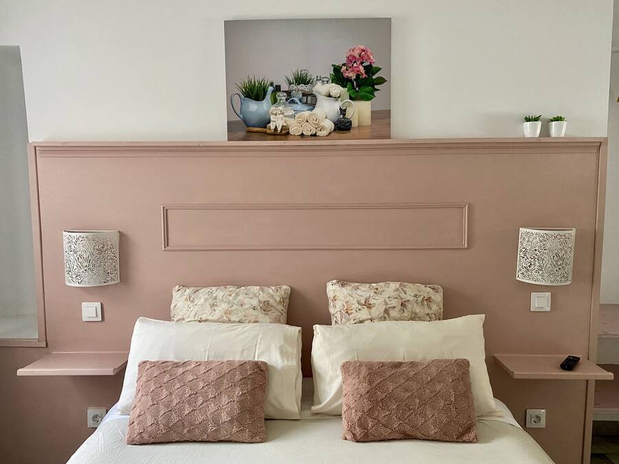Ein Schlafzimmer mit einem Bett, zwei Kissen, einem Nachttisch, einer Wandlampe und einem dekorativen Regal mit Pflanzen und einer Vase.