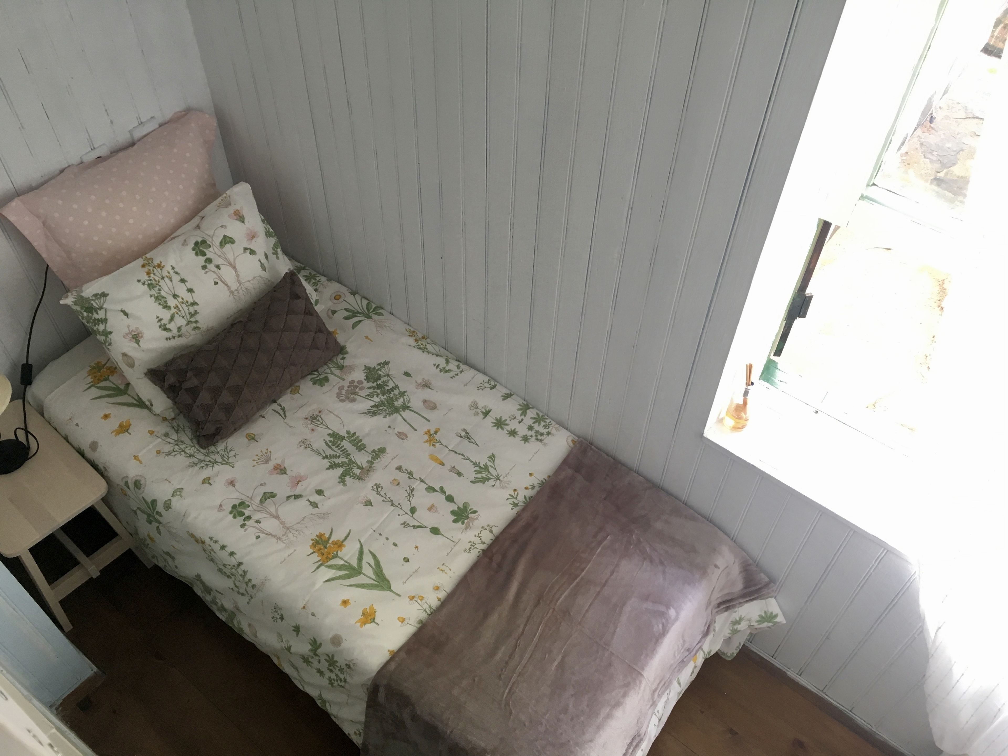 Une petite chambre avec un lit simple, recouverte d’une courtepointe à motifs floraux, un oreiller marron matelassé et un oreiller rose. Il y a une fenêtre sur le côté droit et une petite table avec une lampe sur le côté gauche.