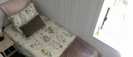Une petite chambre avec un lit simple, recouverte d’une courtepointe à motifs floraux, un oreiller marron matelassé et un oreiller rose. Il y a une fenêtre sur le côté droit et une petite table avec une lampe sur le côté gauche.