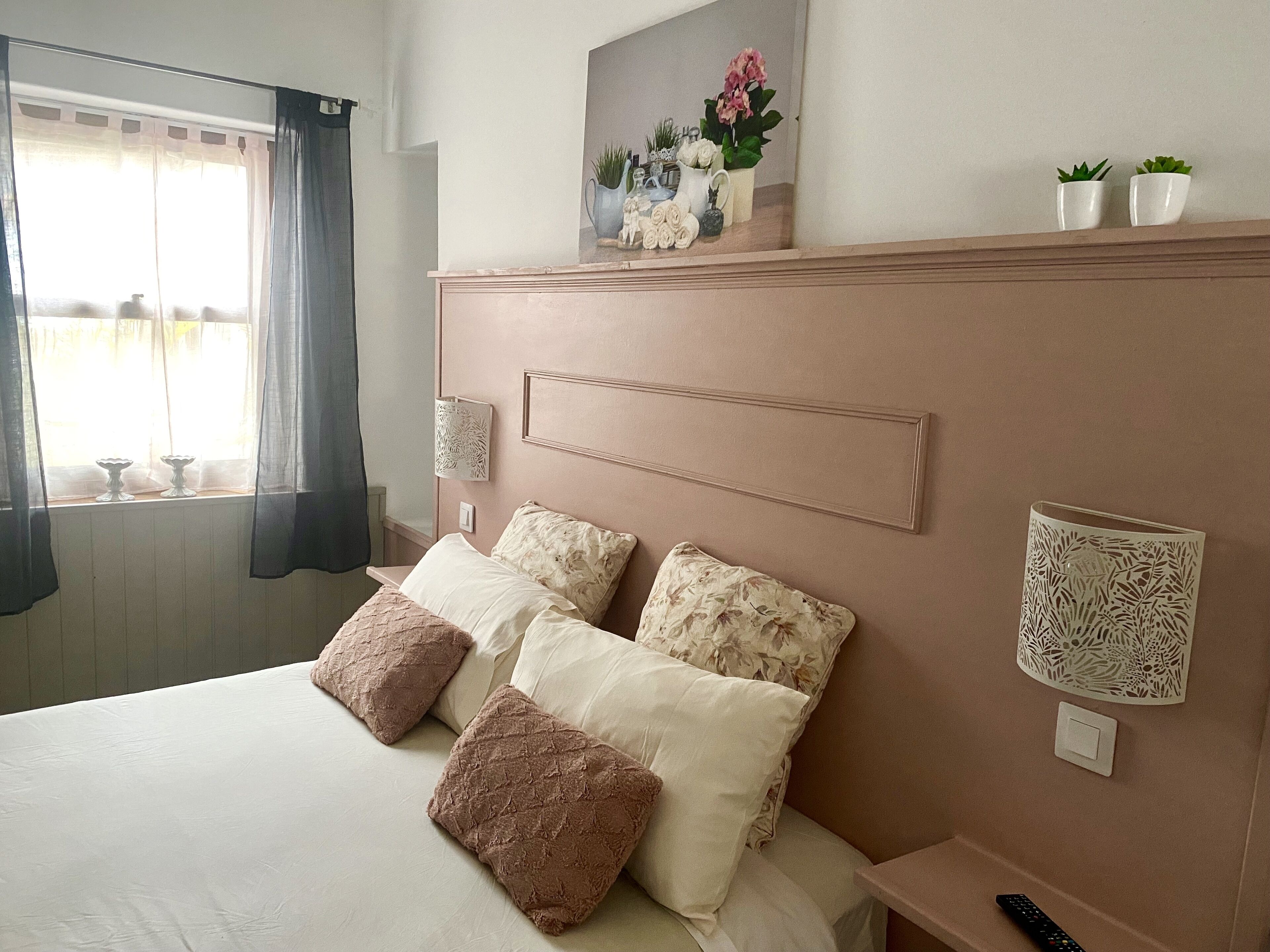 Een netjes opgemaakt bed met vier kussens, een nachtkastje met afstandsbediening, een wandlamp en een raam met dunne gordijnen.