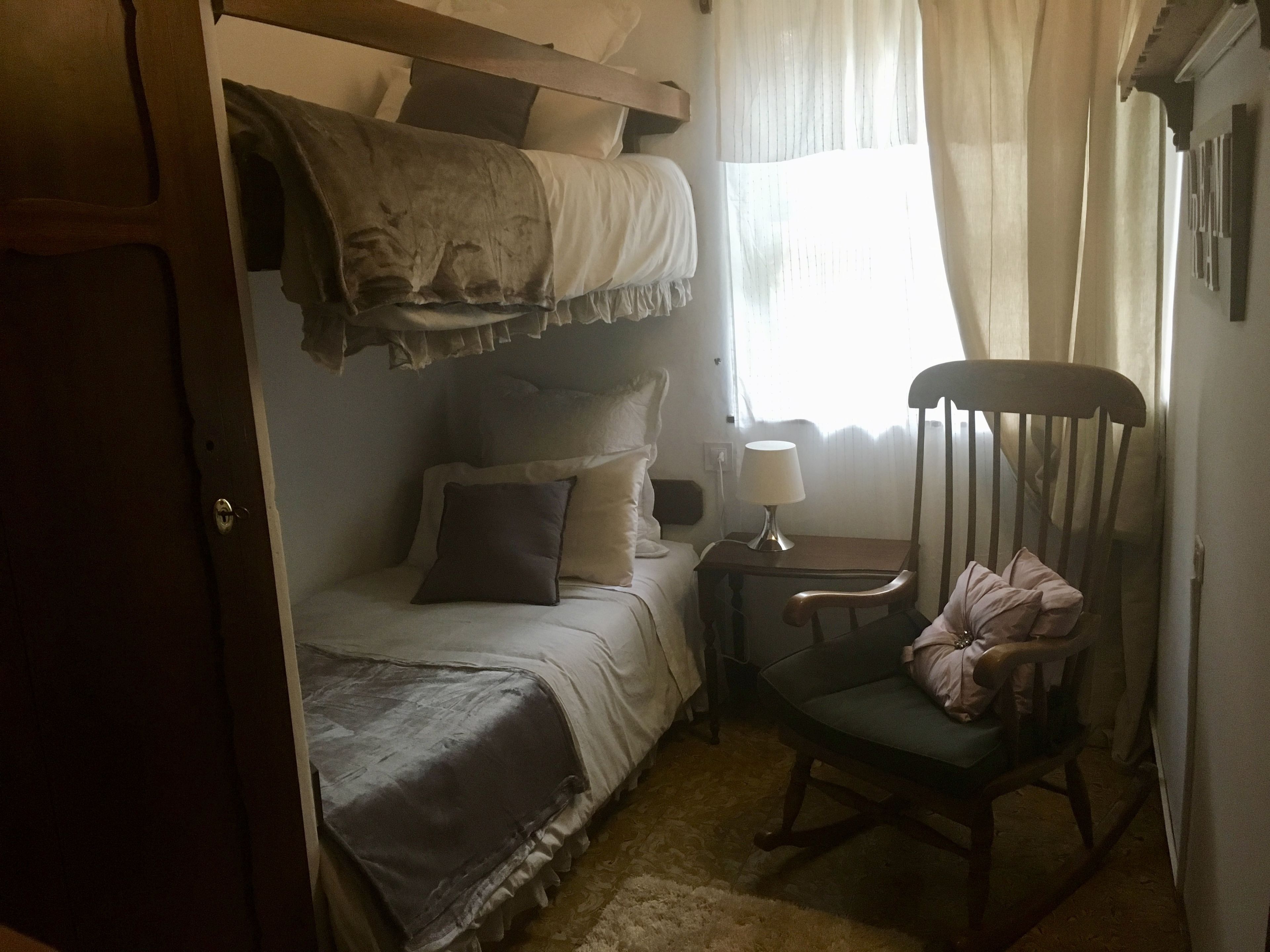 Un dormitorio con una cama litera, una silla, una mesita de noche con lámpara y una ventana con cortinas.