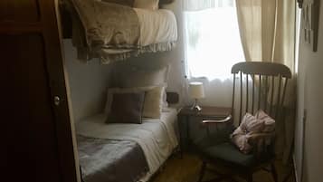 Un dormitorio con una cama litera, una silla, una mesita de noche con lámpara y una ventana con cortinas.