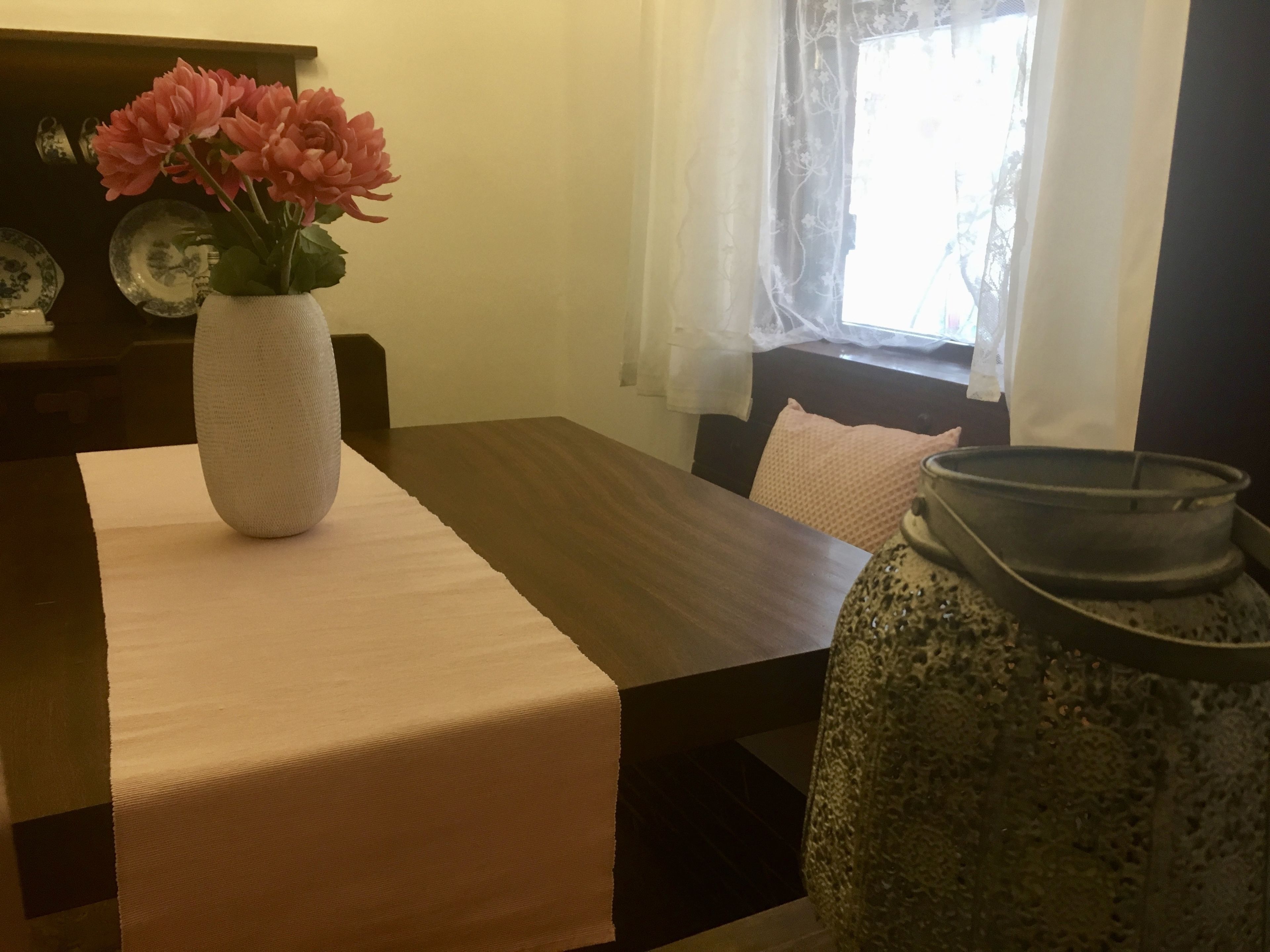 Een eettafel met een vaas roze bloemen, een tafelkleed met textuur en een grote keramieken pot.