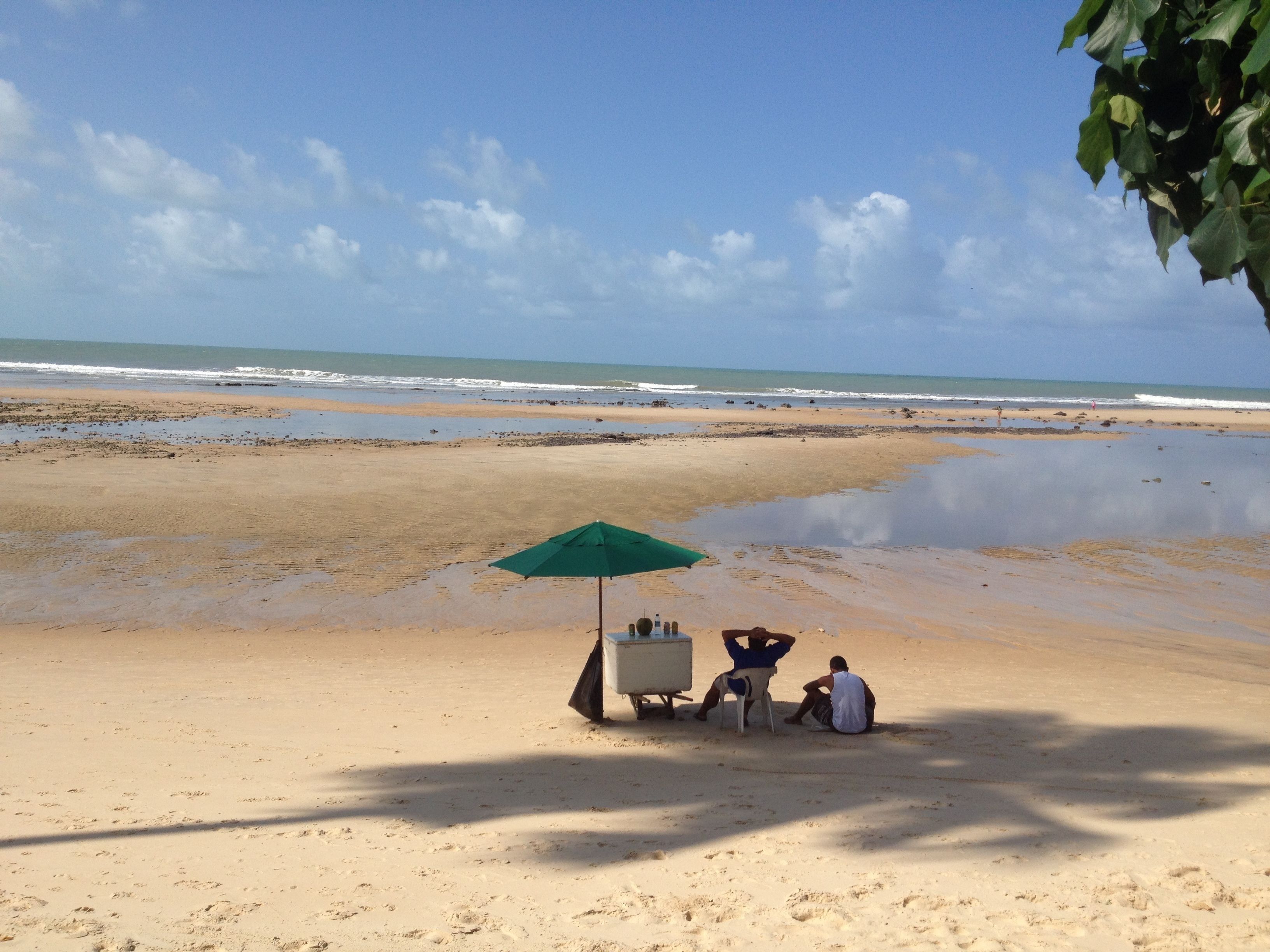 Une plage avec un parasol vert et une petite table, deux personnes assises en dessous, et l’océan en arrière-plan.
