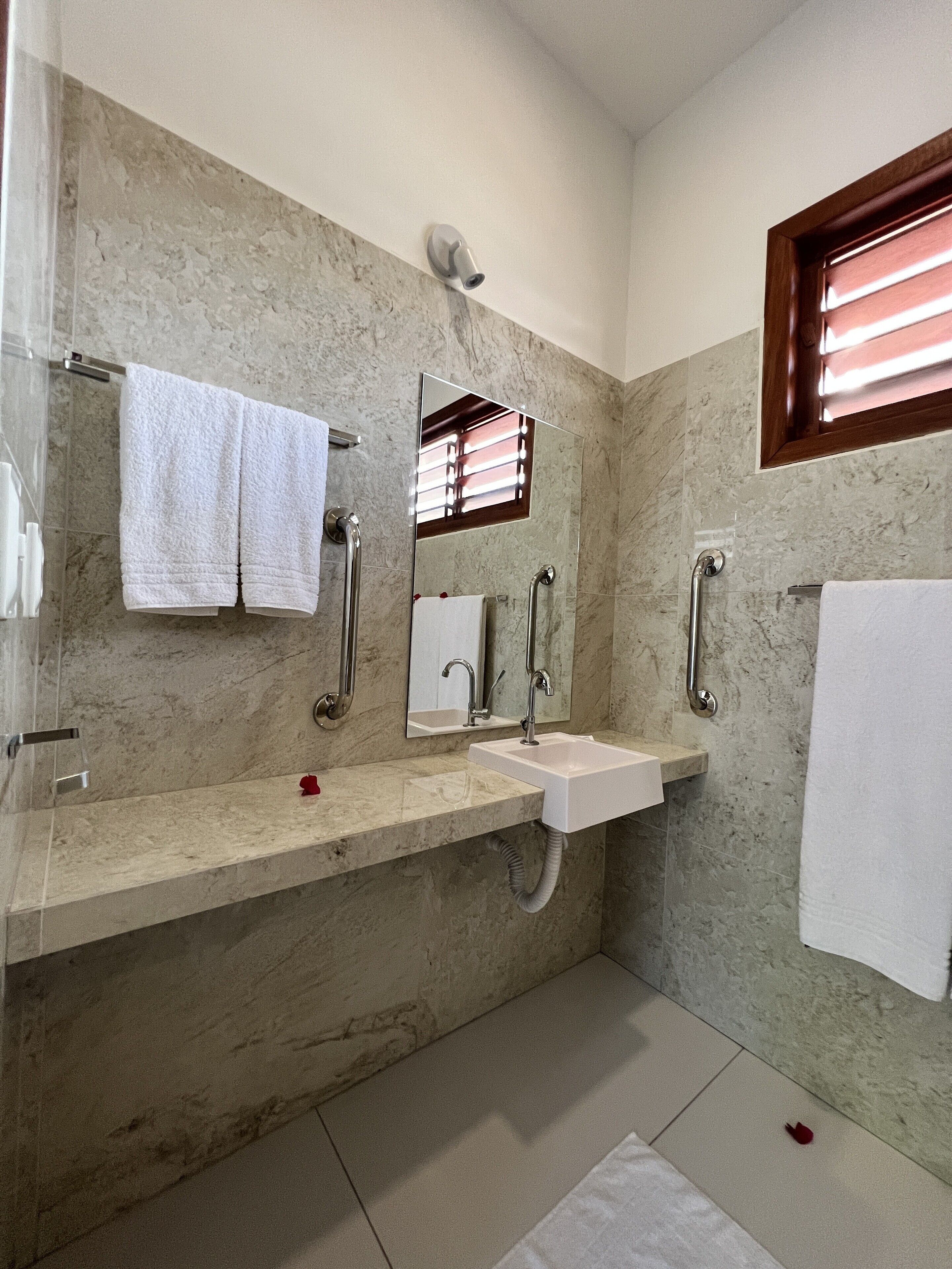 A bathroom with a marble wall, a sink, a mirror, and a window with blinds.