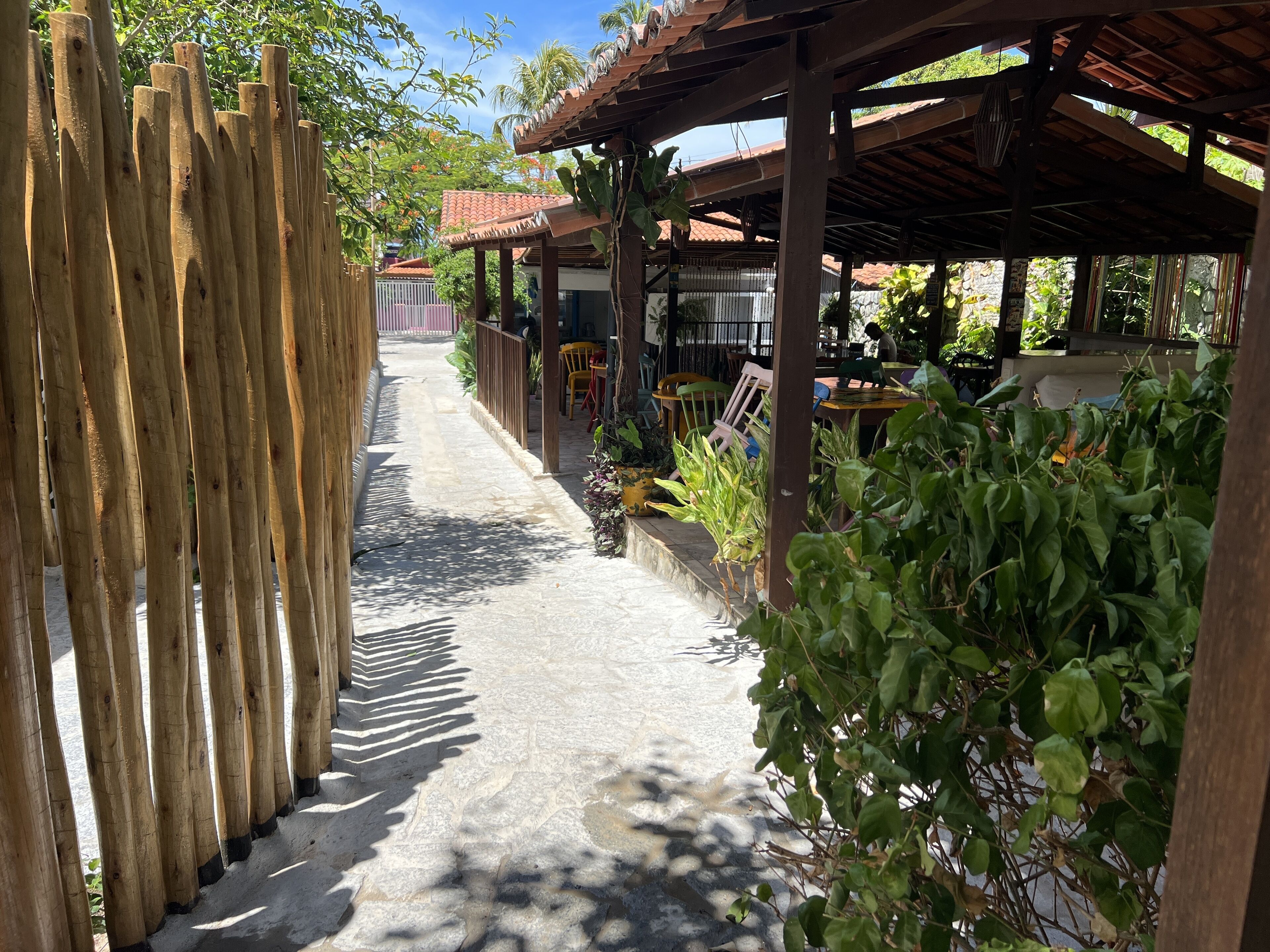 A narrow pathway flanked by wooden fences and lush greenery, leading to a covered outdoor seating area with tables and chairs.