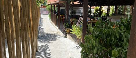 A narrow pathway flanked by wooden fences and lush greenery, leading to a covered outdoor seating area with tables and chairs.