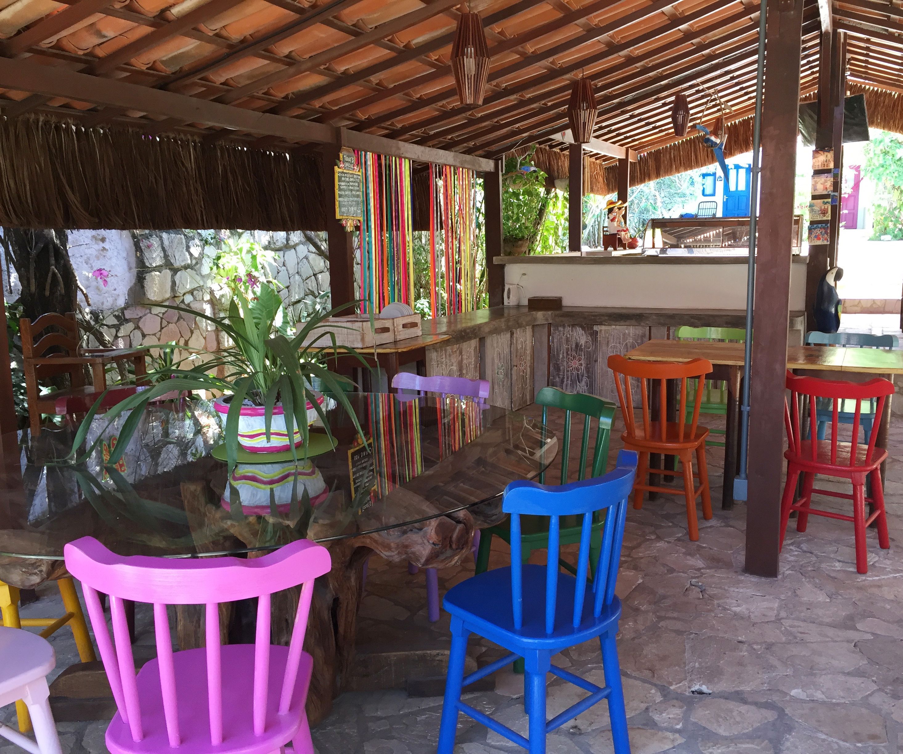 Un espace extérieur abrité, avec des chaises et des tables colorées, un mur en pierre et un toit de chaume.