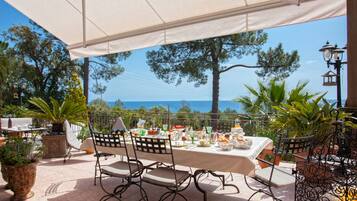 An outdoor dining area with a table set for a meal, surrounded by chairs and potted plants, overlooking a scenic view of the sea.
