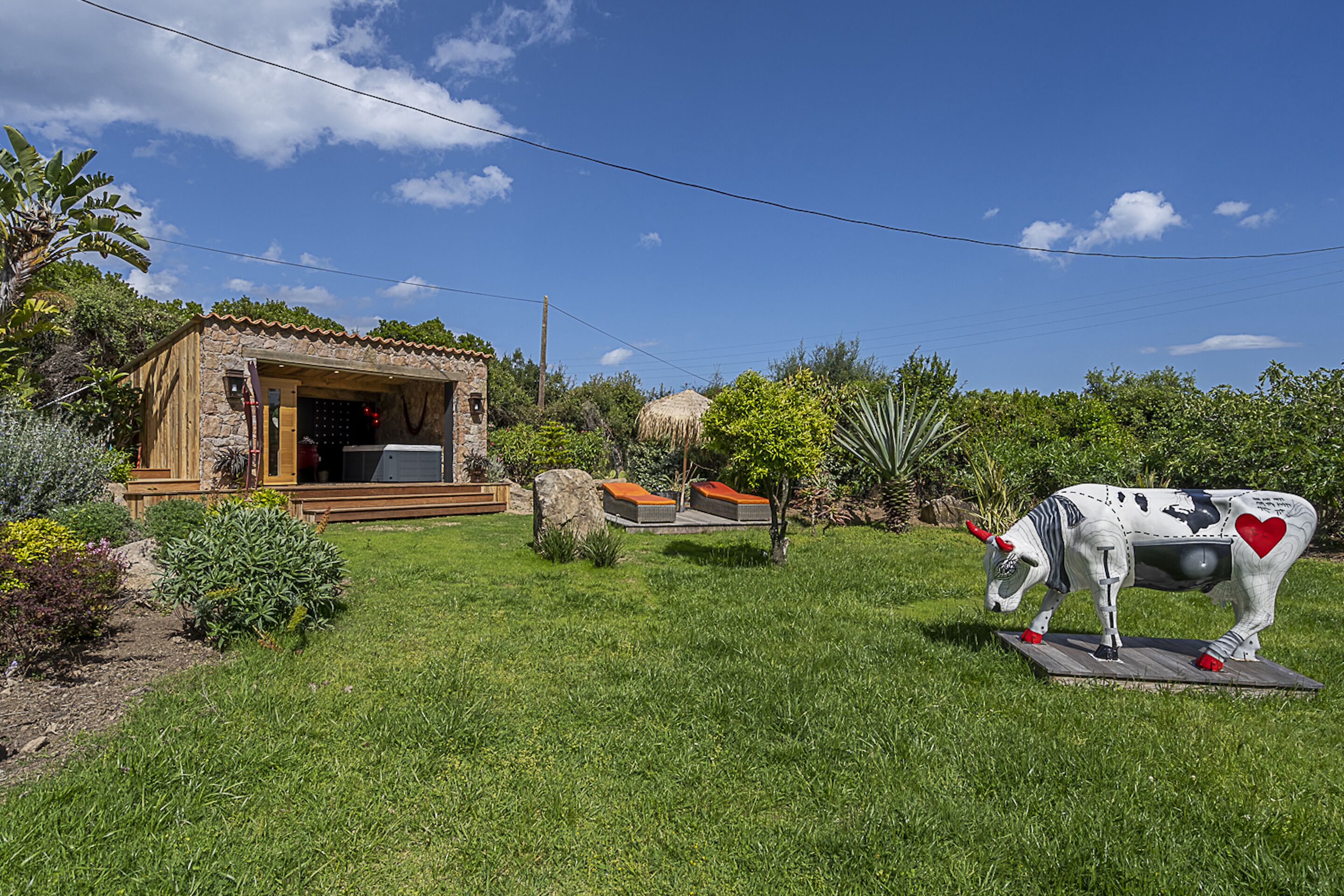 A rustic house with a wooden exterior and a stone foundation, surrounded by a well-maintained garden with various plants and a decorative cow statue.