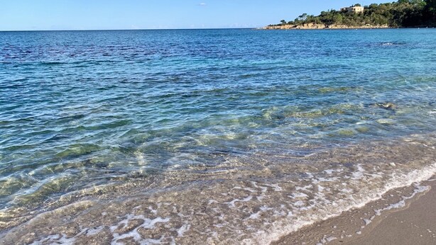 A beach with clear water, a sandy shore, and a distant shoreline with buildings. - Casa Del Sole Chambre d'hôtes de Charme (Sari-Solenzara)