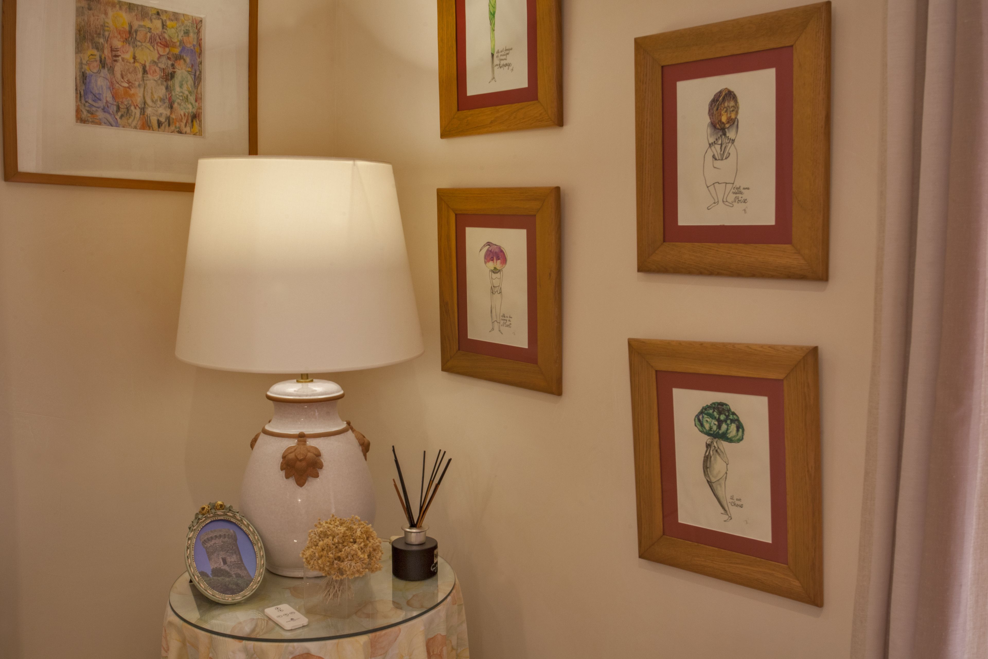 A room with a lamp on a glass table, surrounded by framed artworks on the wall.