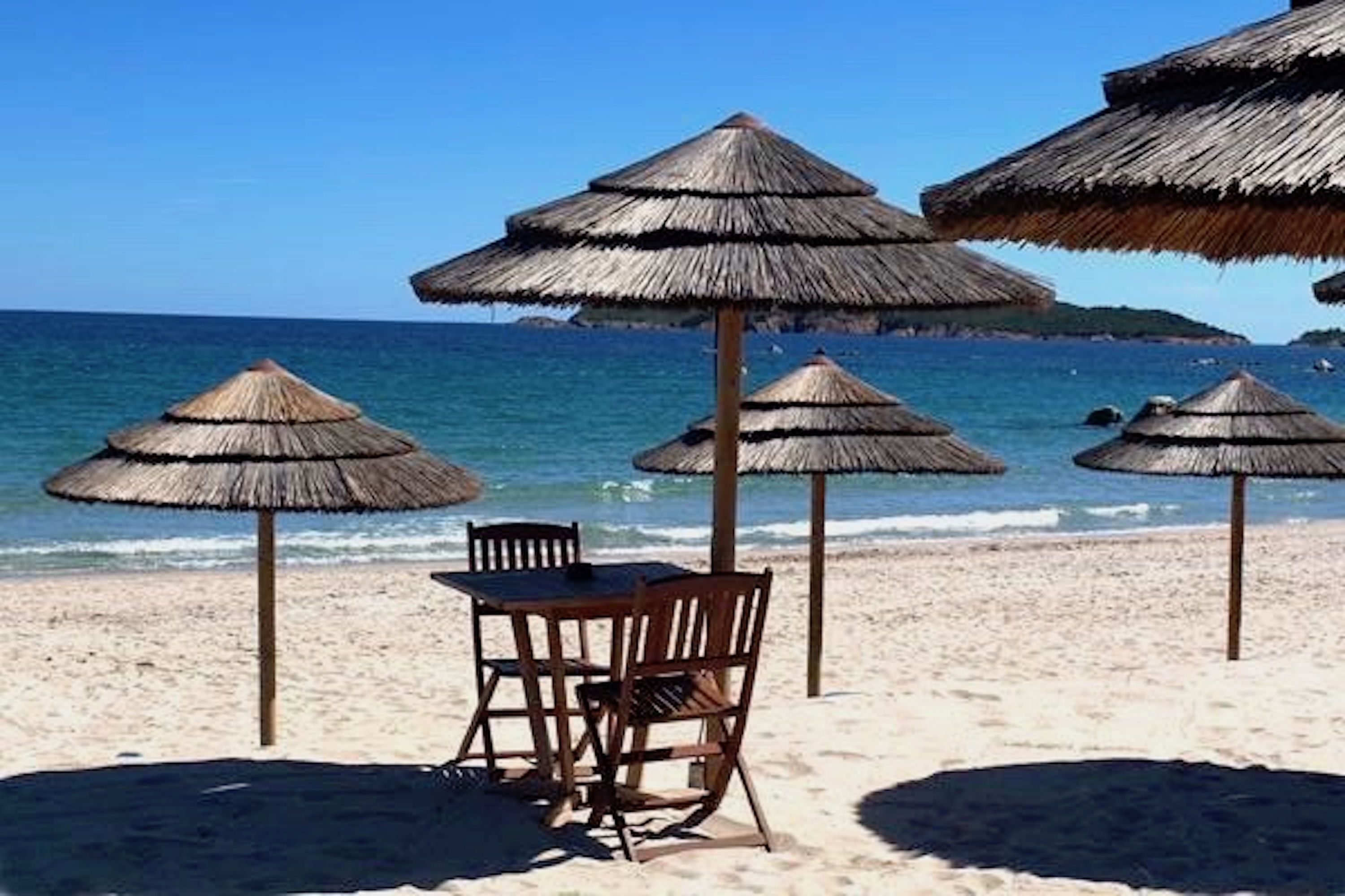 A beach with thatched umbrellas and a table with chairs.