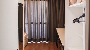 Un pasillo estrecho con suelo de madera, una puerta con placa de número, una mesa con sillas y un estante con zapatos.