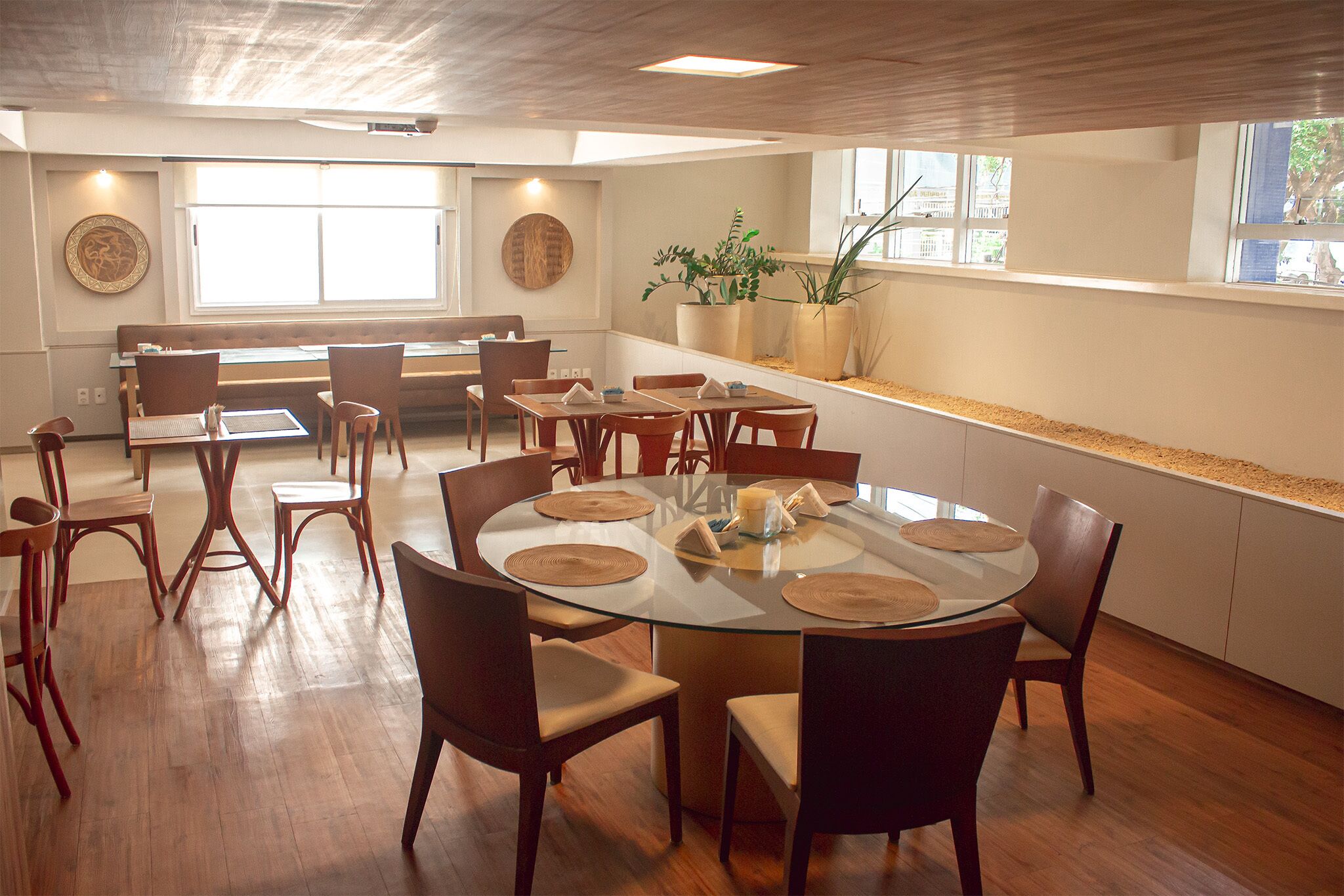 Un espace repas avec une table ronde en verre entourée de chaises en bois, un banc le long du mur et de grandes fenêtres laissant entrer la lumière naturelle.