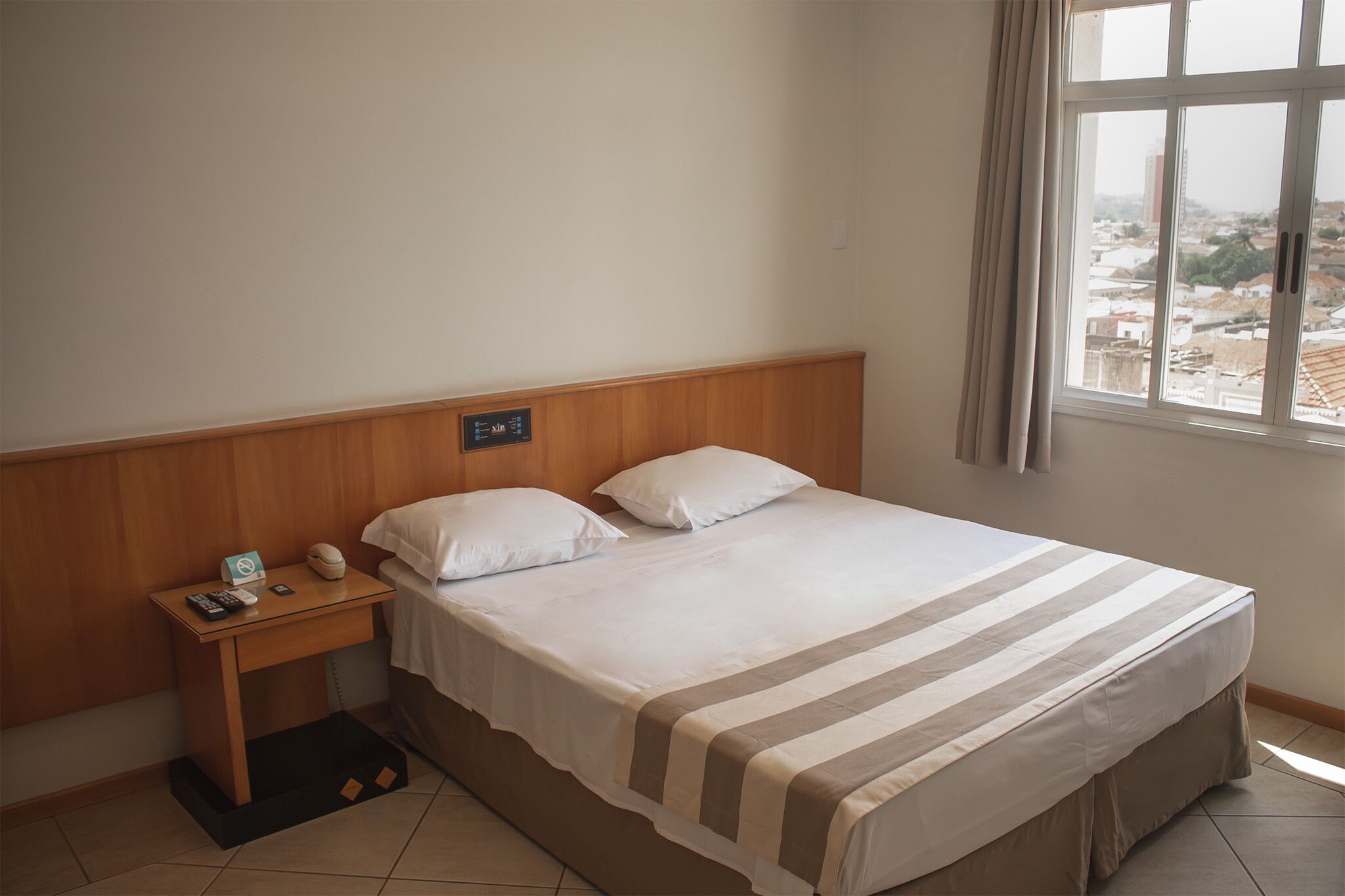 A hotel room with a bed, a nightstand, a window with a view of the city, and a wooden headboard.