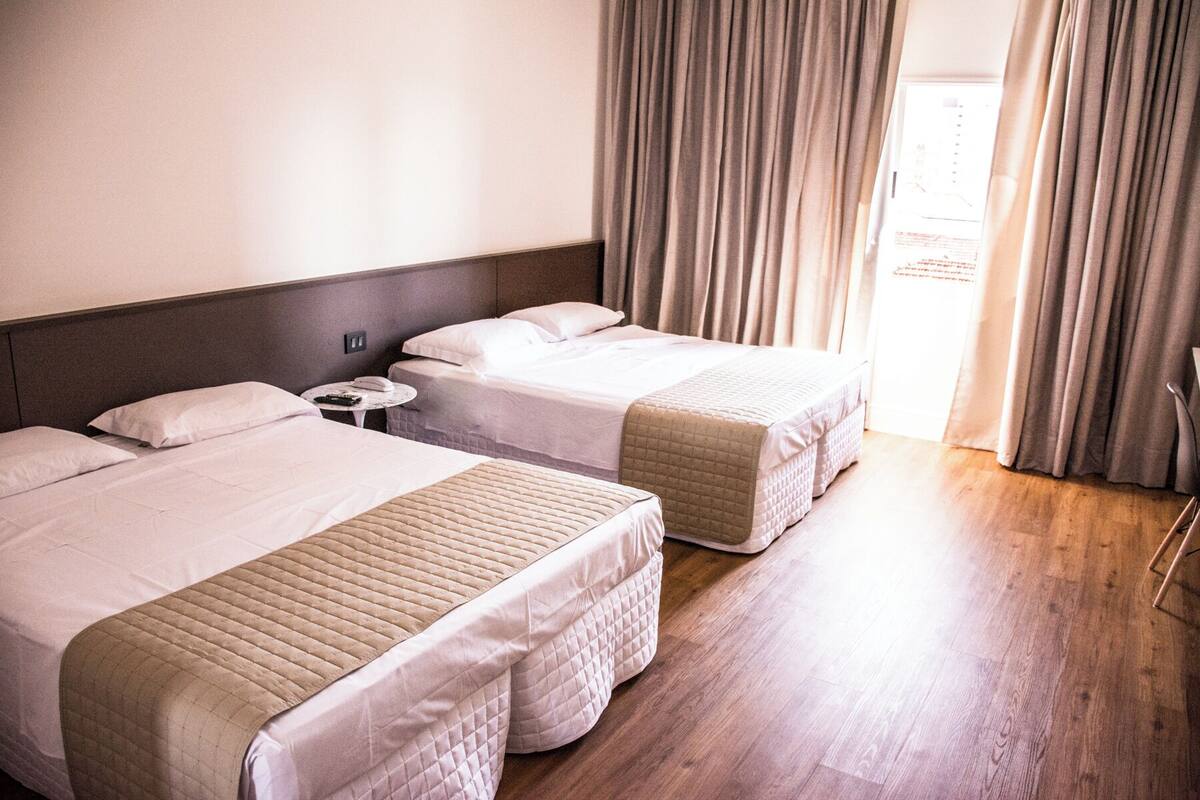 A hotel room with two beds, a small table, and a view of the outside through curtains.