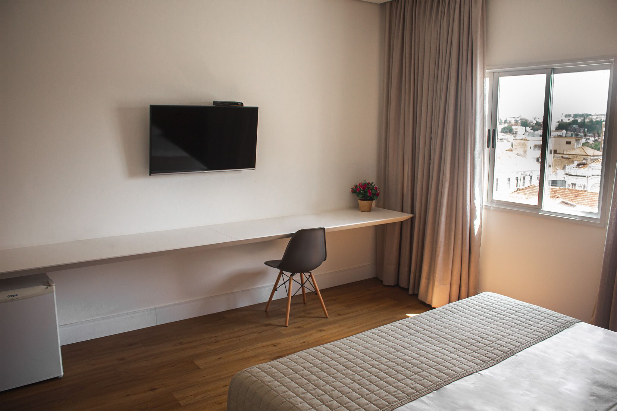 Habitación de hotel con cama, escritorio con silla, televisor montado en la pared y ventana con cortinas.