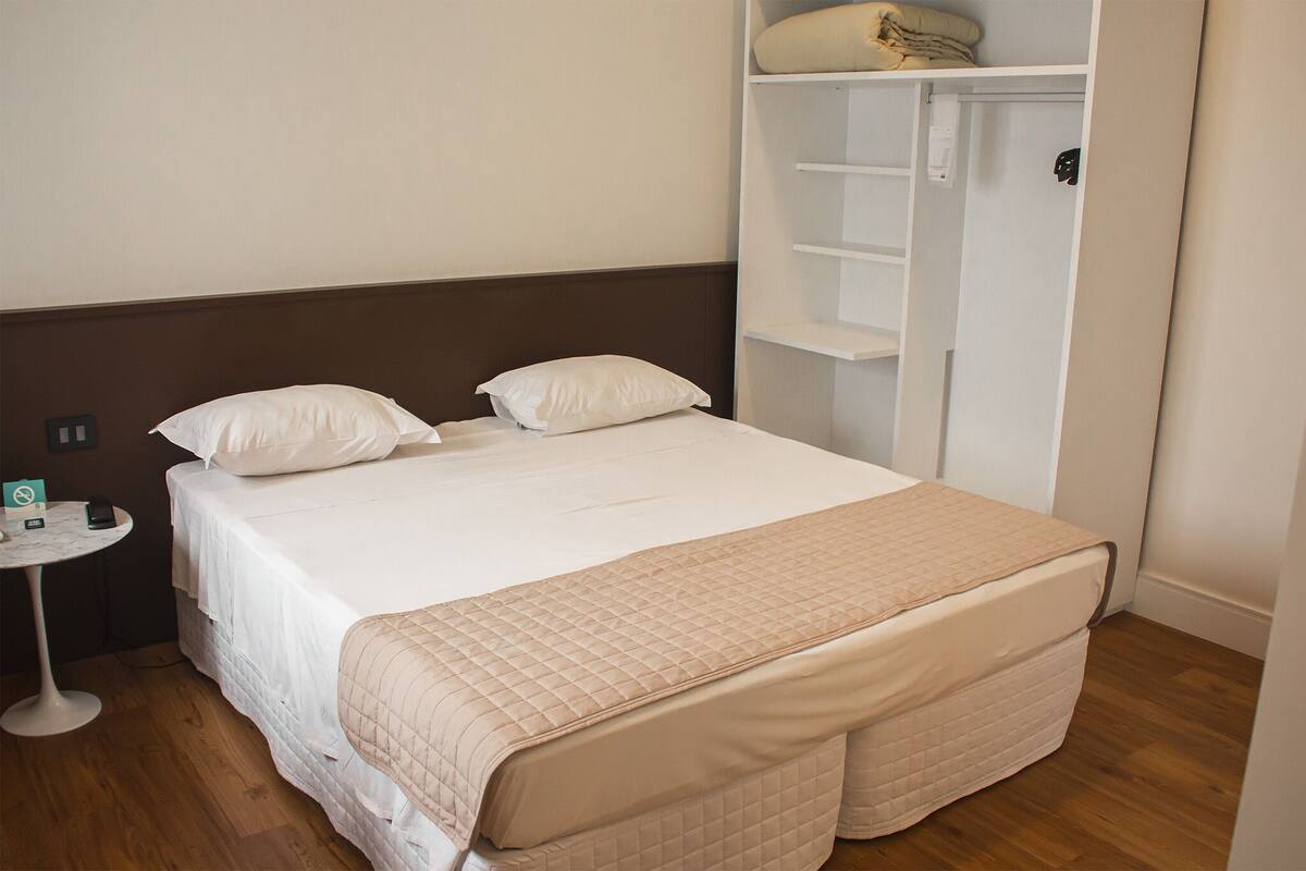 A neatly made bed with a quilted cover, a bedside table with a phone and a card, and a white wardrobe with shelves.