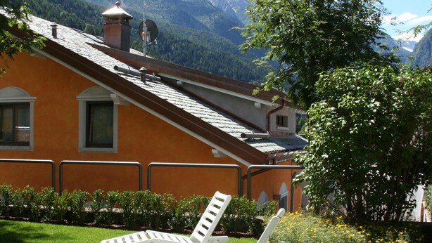 Una casa con techo inclinado, una estructura similar a una torre y un jardín con césped y dos sillones blancos.