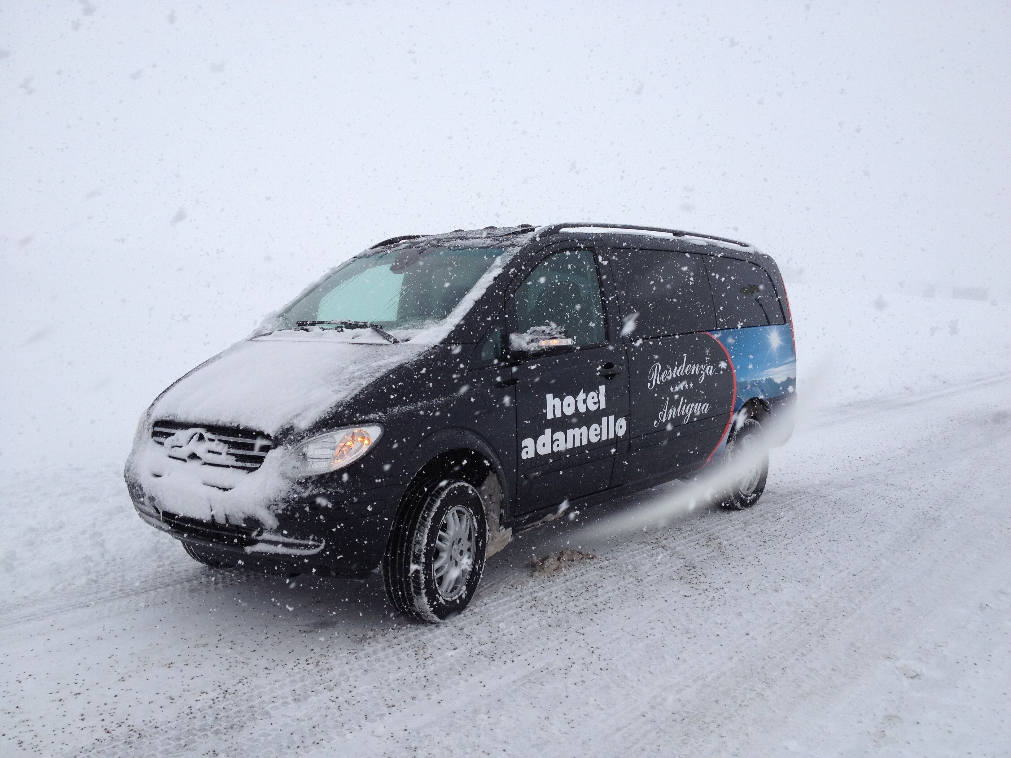 A black van with "hotel adamello" and "Residencia Antigua" on its side, driving on a snowy road.
