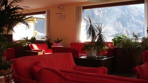 Habitación de hotel con muebles rojos, amplias ventanas con vistas a la montaña y varias plantas en macetas.