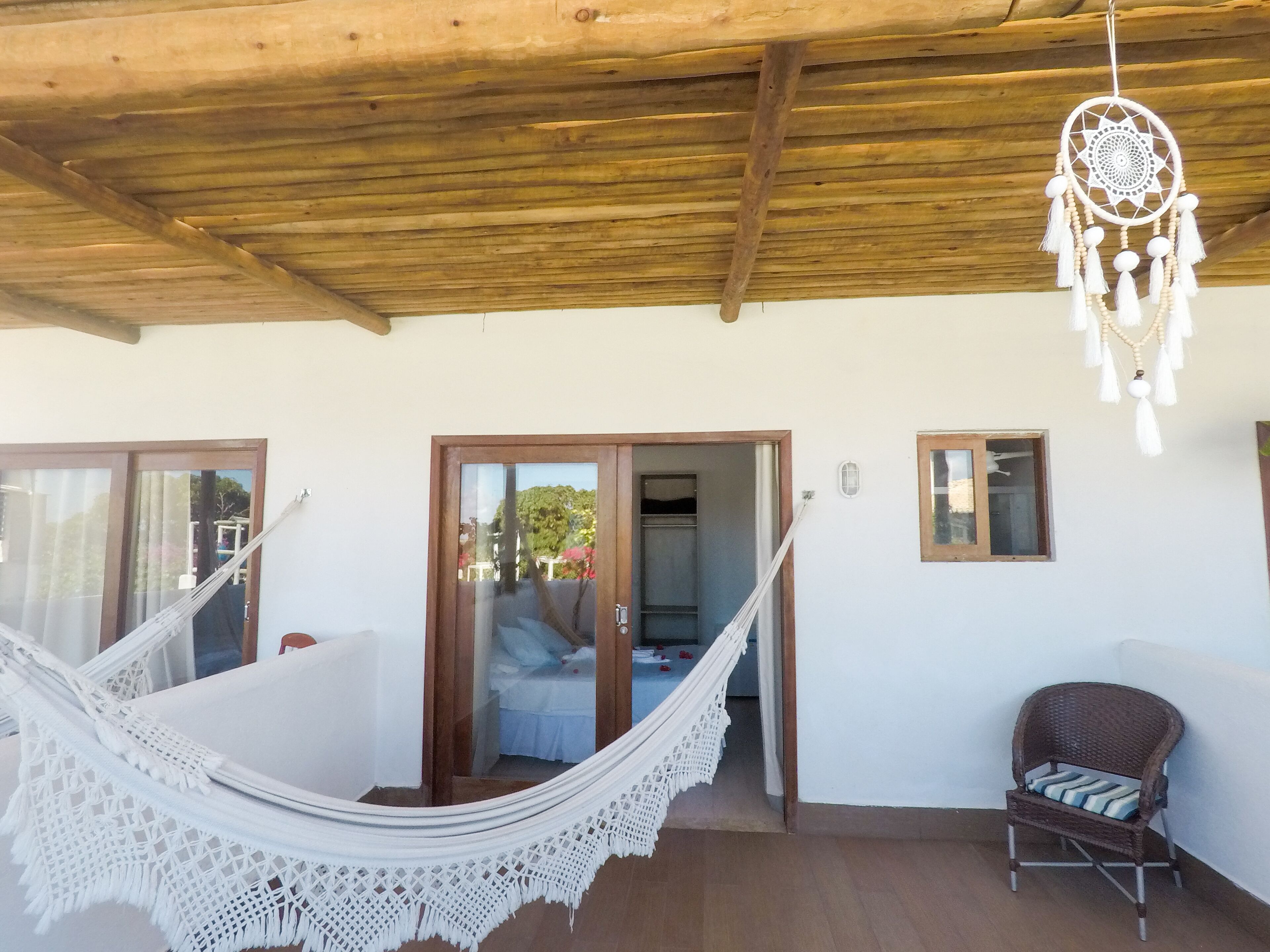 A room with a hammock, a wicker chair, a dreamcatcher, and a view of a garden through a door.