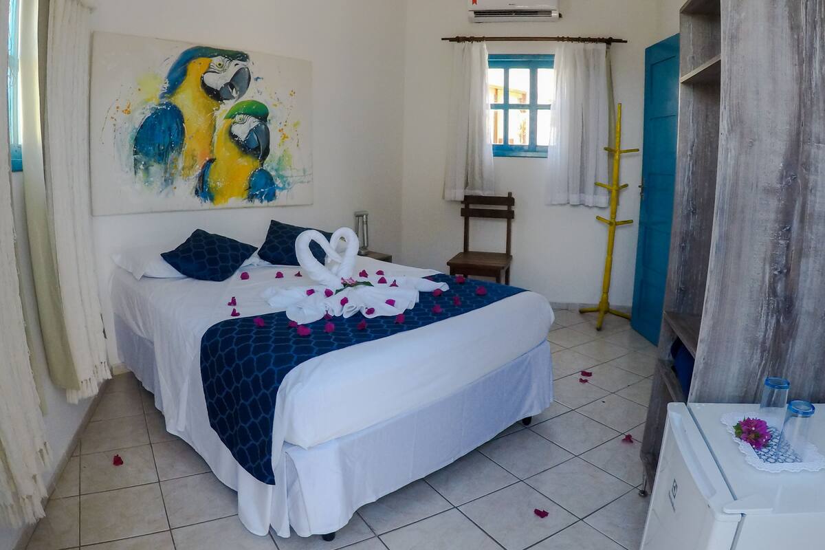Quarto de hotel com cama, pintura, janela com cortinas, cadeira e porta de madeira.