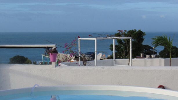 En pool med krystalklart havudsyn, en hvid bygning med pergola, og en pink blomsterpotte.