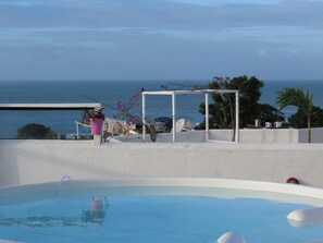 A pool with a clear view of the ocean, a white structure with a pergola, and a pink flower pot. - Pousada Mediterrânea (Tibau do Sul)
