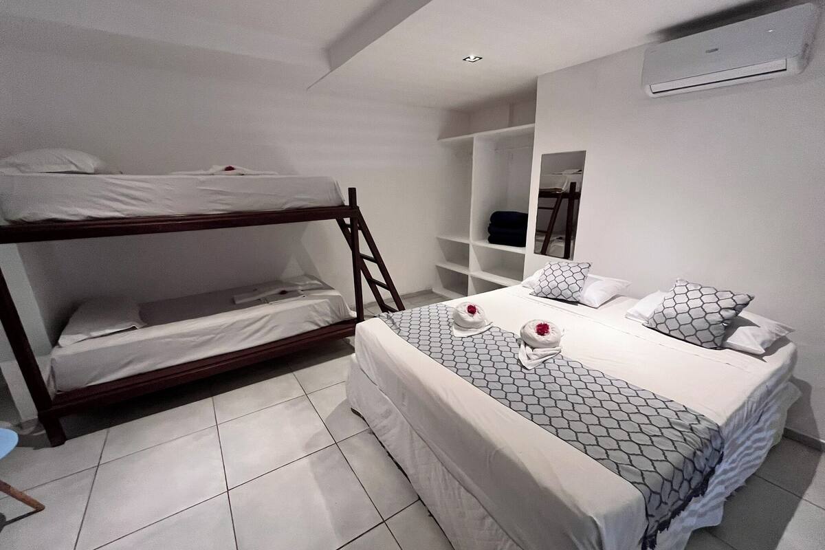 Quarto de hotel com beliche, cama de casal com roupa de cama estampada, guarda-roupa e espelho.
