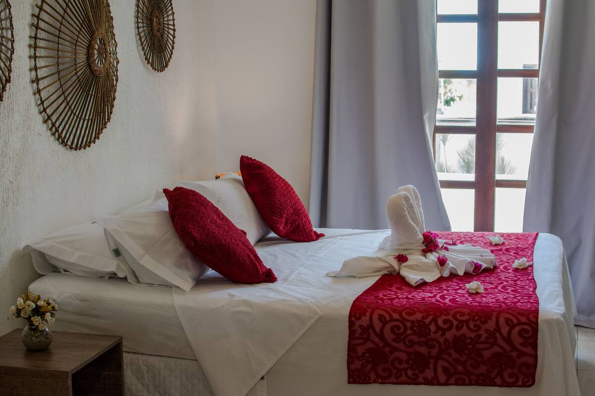Uma cama bem arrumada com roupa de cama branca e vermelha, um vaso de flores em uma mesa de cabeceira e uma janela com cortinas.